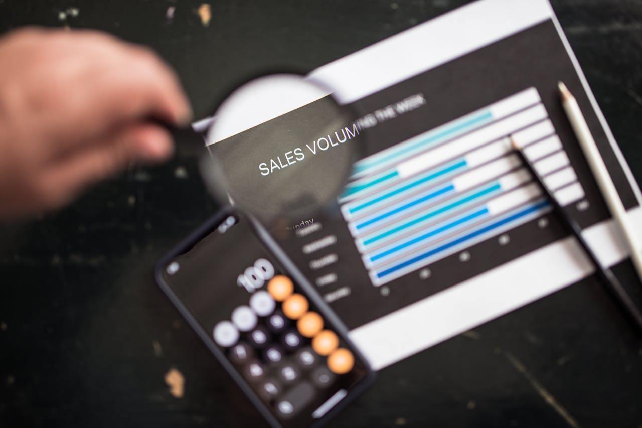 Close-up of a magnifying glass focusing on a sales volume chart next to a smartphone calculator app.